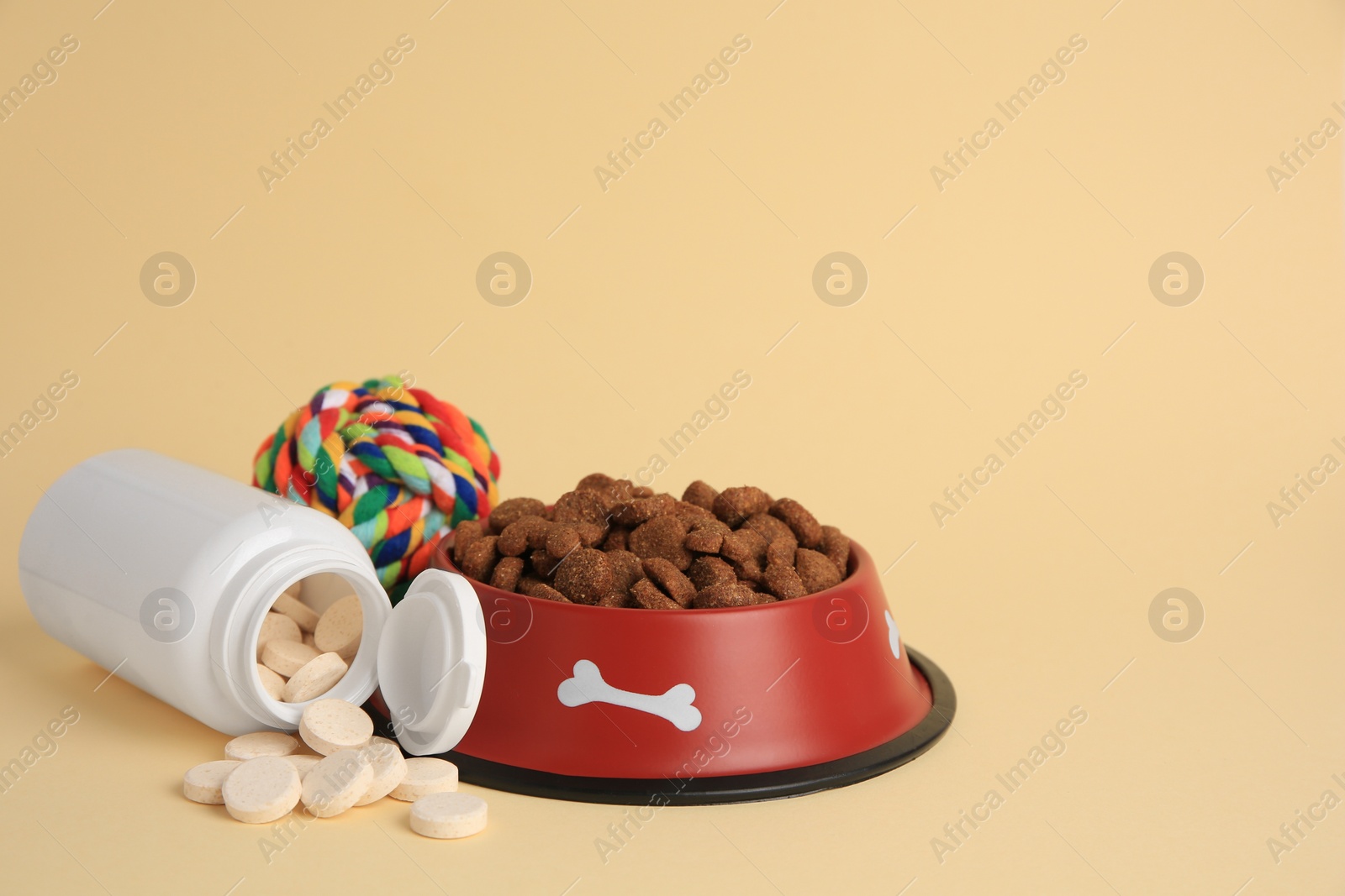 Photo of Bottle with vitamins, dry pet food in bowl and toy on beige background, space for text