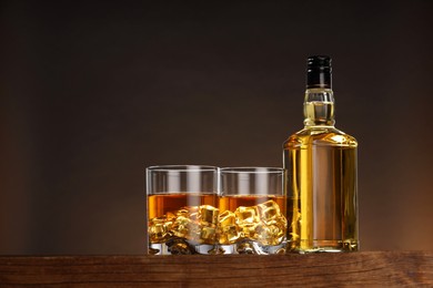Photo of Whiskey with ice cubes in glasses and bottle on wooden table against brown background, space for text