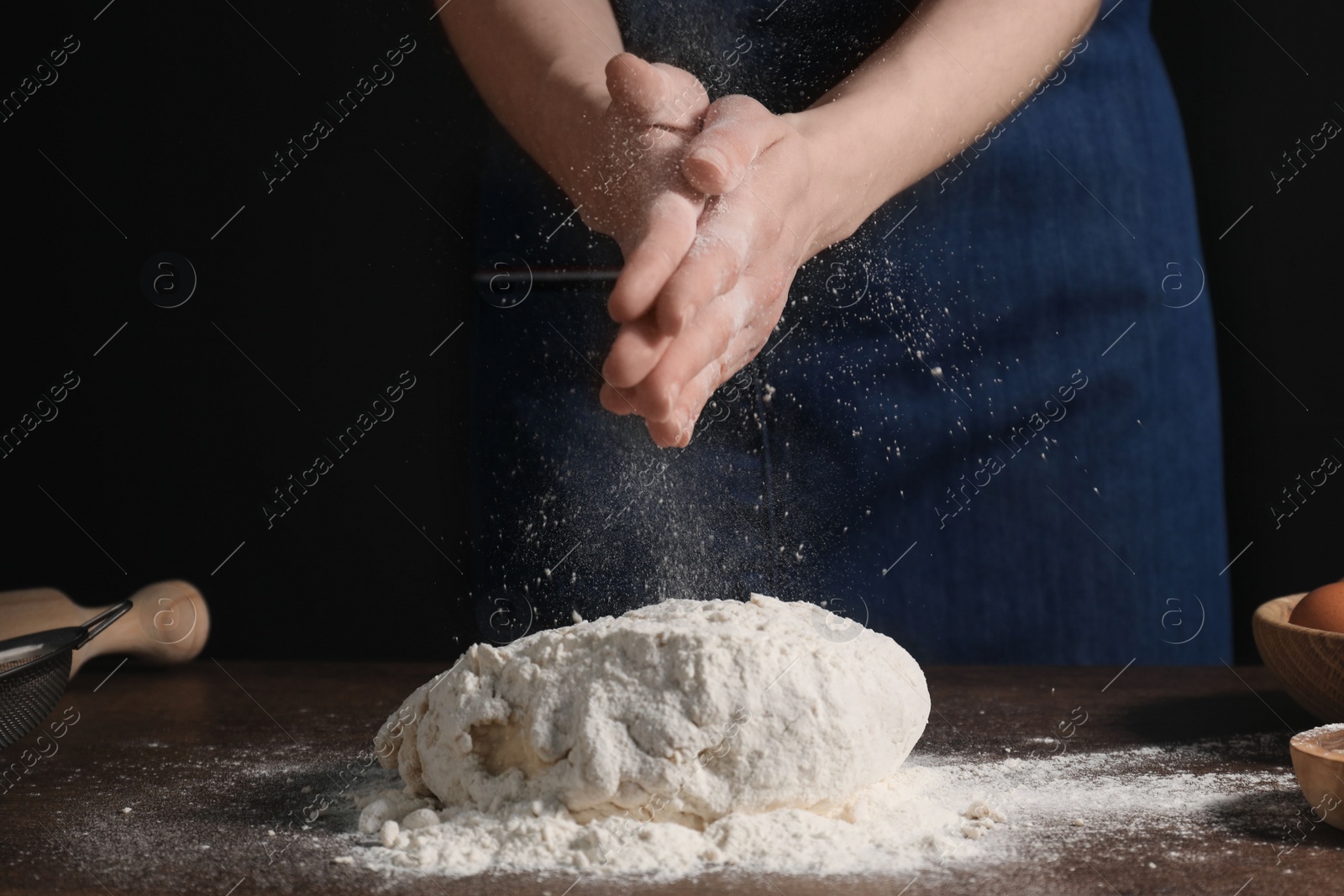 Photo of Making bread. Woman sprinkling flour over dough at table on dark background, closeup