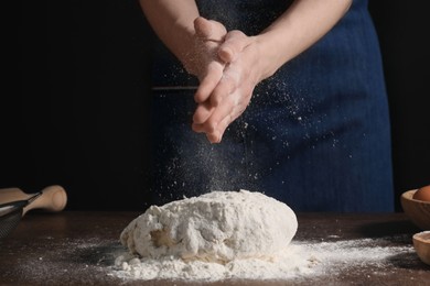 Making bread. Woman sprinkling flour over dough at table on dark background, closeup