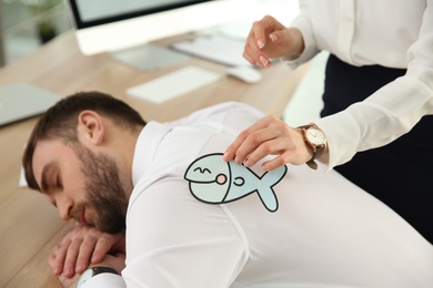 Photo of Young woman sticking paper fish to colleague's back while he sleeping in office, closeup. Funny joke