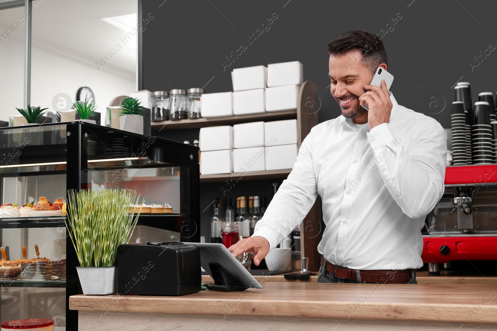 Photo of Business owner in his cafe. Man talking on phone while using tablet near showcase with pastries