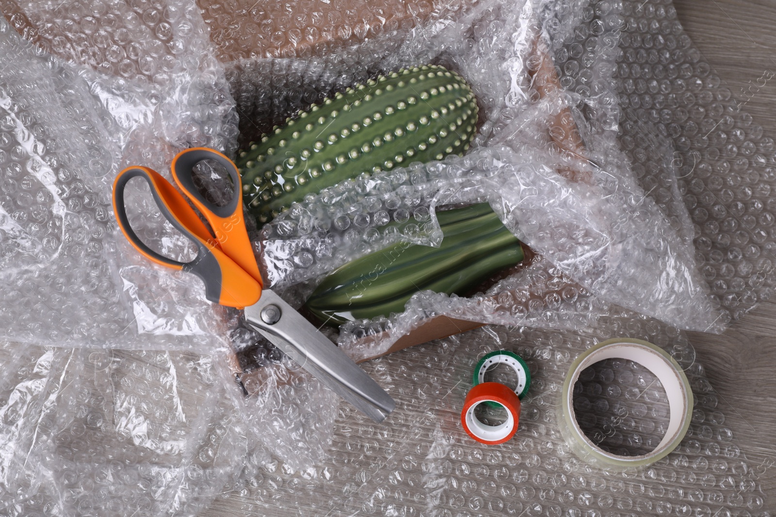 Photo of Ceramic cacti with bubble wrap in cardboard box, scissors and adhesive tapes on wooden table, above view