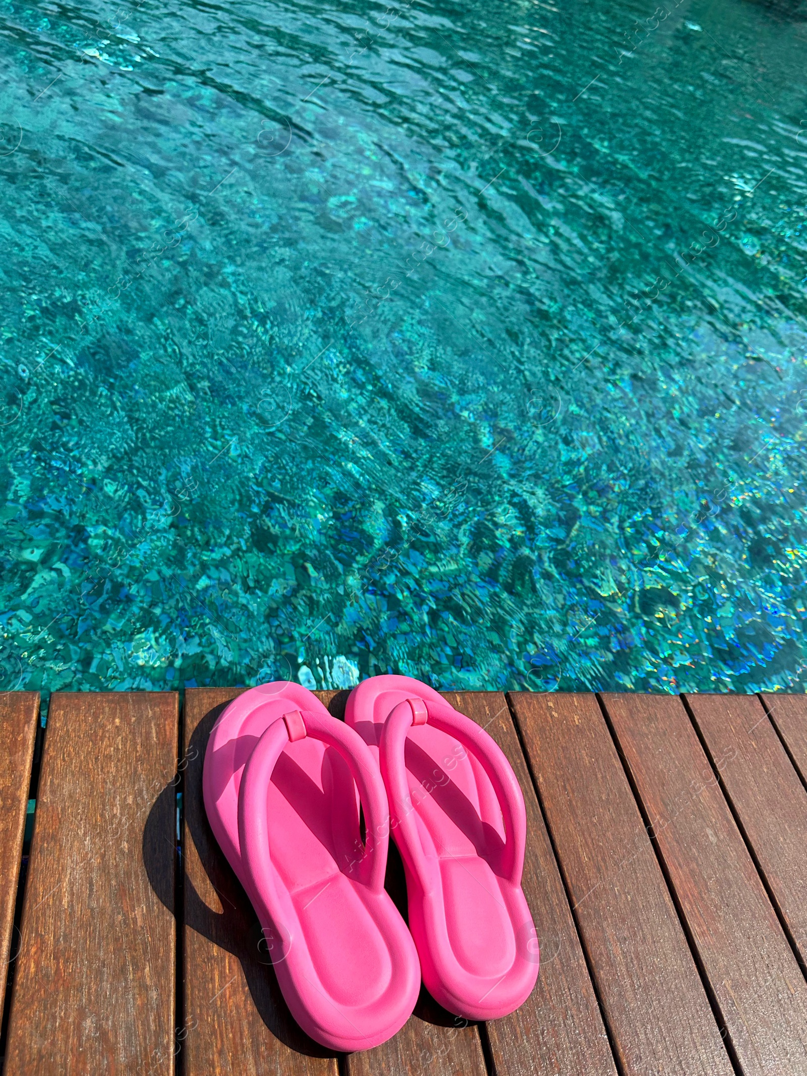 Photo of Clear rippled water in swimming pool and pink flip-flops on wooden deck outdoors. Space for text