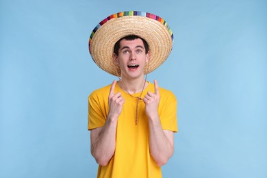 Young man in Mexican sombrero hat pointing at something on light blue background