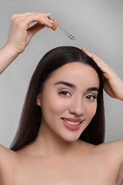 Happy young woman applying essential oil onto hair roots on light grey background