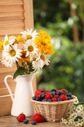 Wicker bowl with different fresh ripe berries and beautiful flowers on wooden table outdoors