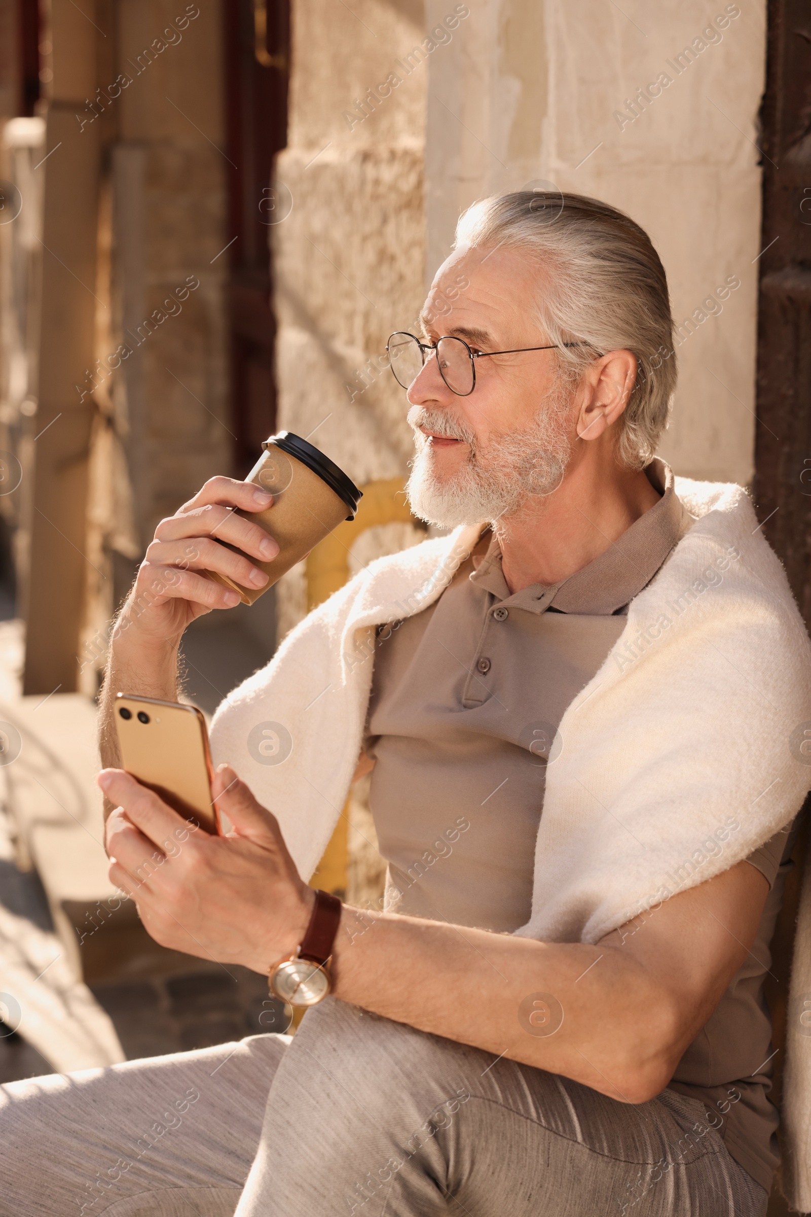 Photo of Handsome senior man sitting on doorstep with smartphone and drinking coffee outdoors