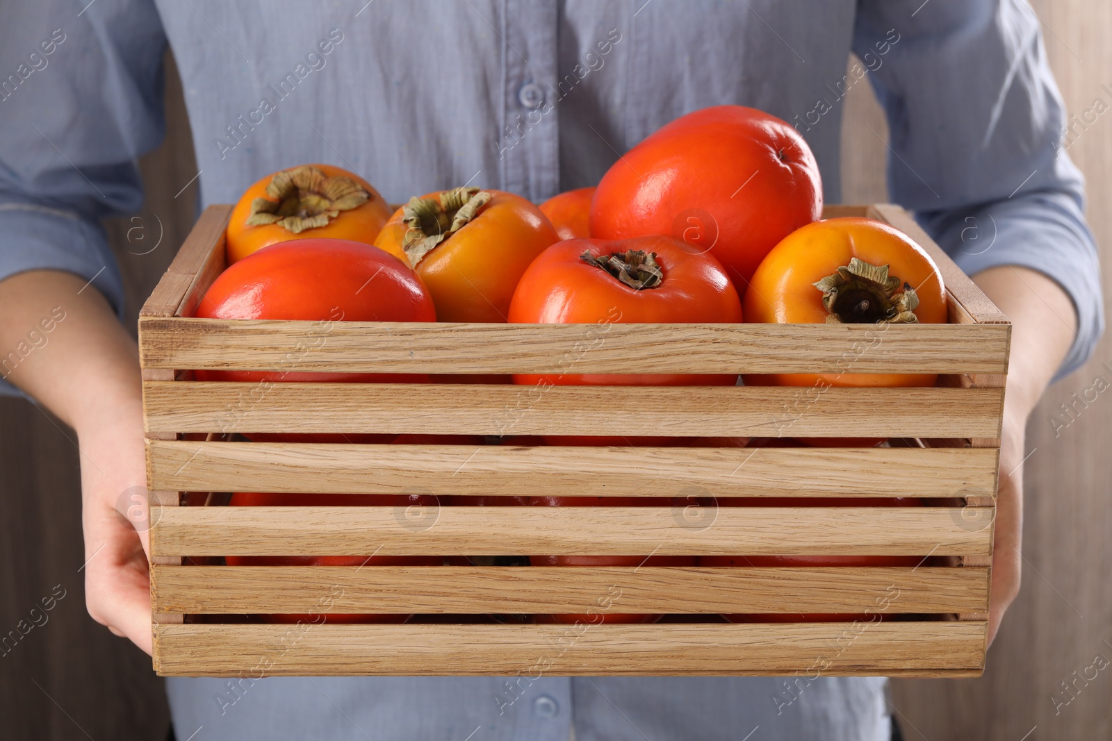 Photo of Woman holding wooden crate with delicious ripe juicy persimmons on blurred background, closeup