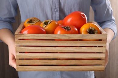 Photo of Woman holding wooden crate with delicious ripe juicy persimmons on blurred background, closeup