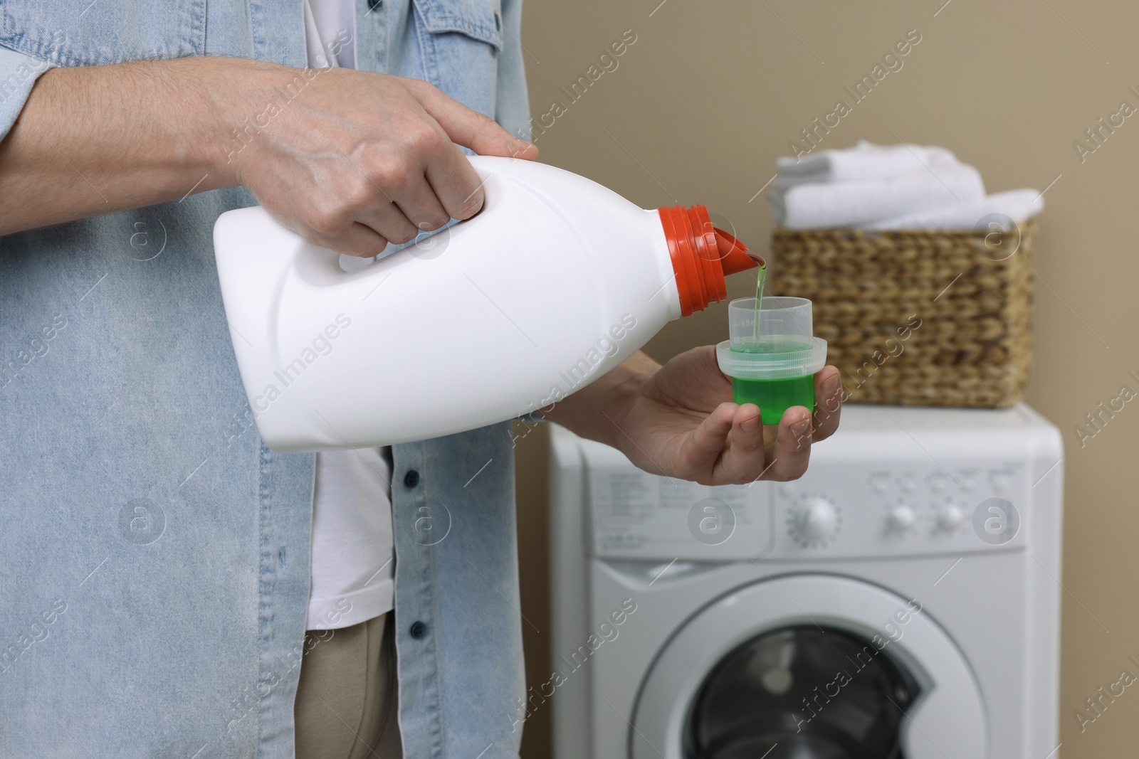 Photo of Man pouring fabric softener from bottle into cap near washing machine indoors, closeup