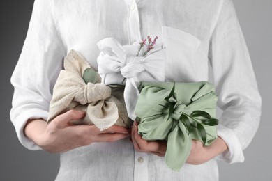 Photo of Furoshiki technique. Woman holding gifts packed in different fabrics decorated with plants on gray background, closeup