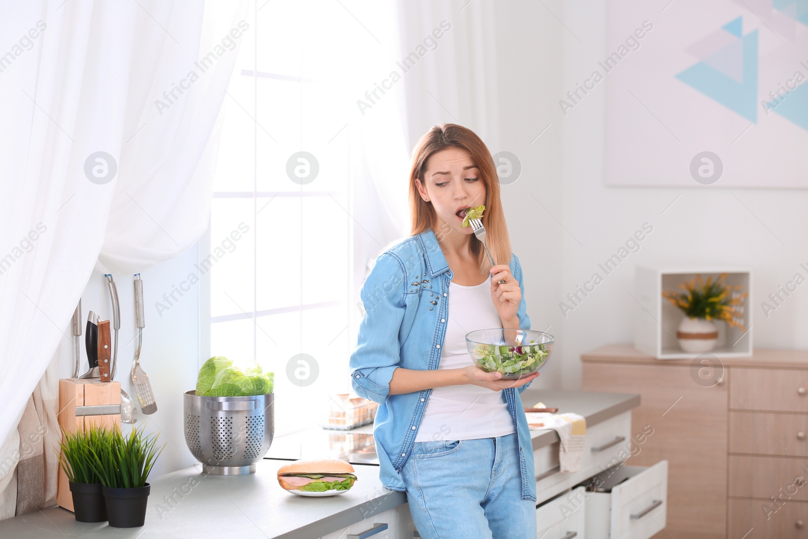 Photo of Emotional young woman eating salad instead of sandwich in kitchen. Healthy diet