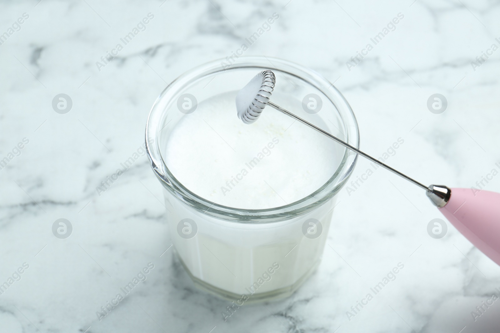 Photo of Mini mixer (milk frother) and whipped milk in glass on white marble table, closeup