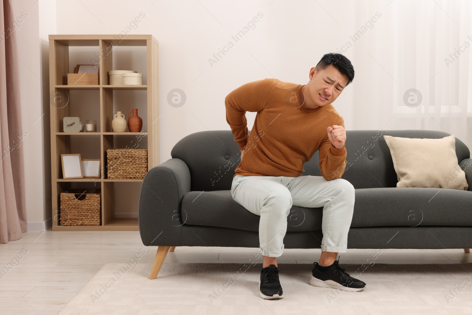 Photo of Asian man suffering from back pain on sofa indoors