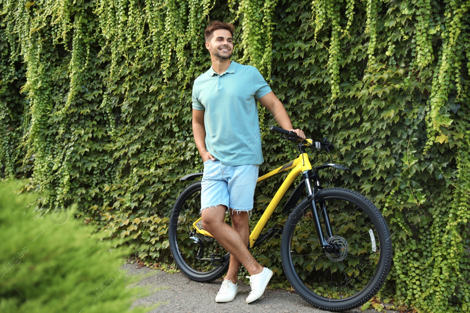 Photo of Handsome young man with bicycle near wall covered with green ivy vines on city street