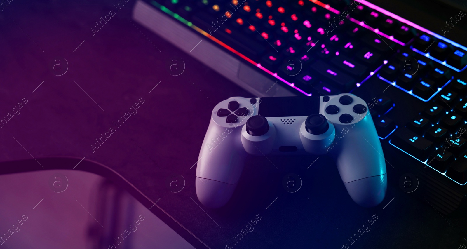 Photo of Playing video games. Computer keyboard with RGB lighting and wireless controller on table indoors