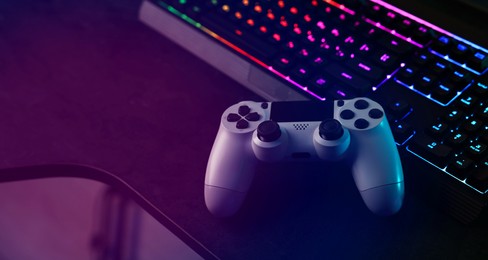 Playing video games. Computer keyboard with RGB lighting and wireless controller on table indoors