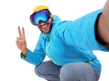Smiling young man in ski goggles taking selfie and showing peace sign on white background