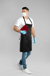 Photo of Waiter in medical face mask with menu on light grey background