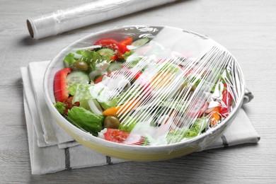 Photo of Bowl of fresh salad with plastic food wrap on white wooden table, closeup