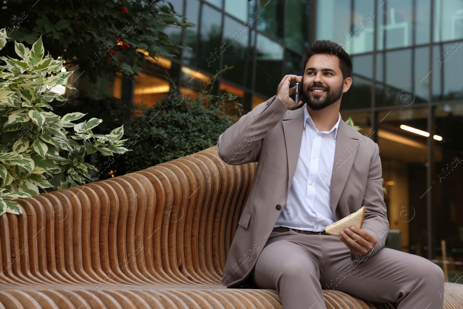 Photo of Lunch time. Young businessman with sandwich talking on smartphone on bench outdoors