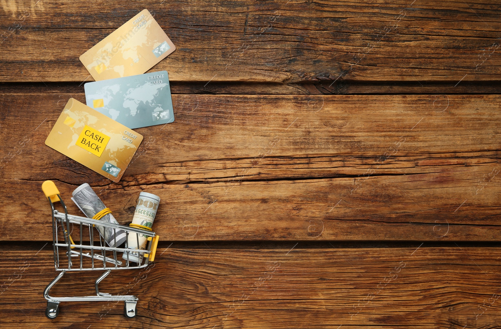 Photo of Credit cards and rolled dollar banknotes in shopping cart on wooden background, flat lay with space for text. Cashback concept