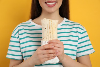 Happy young woman holding tasty shawarma on yellow background, closeup