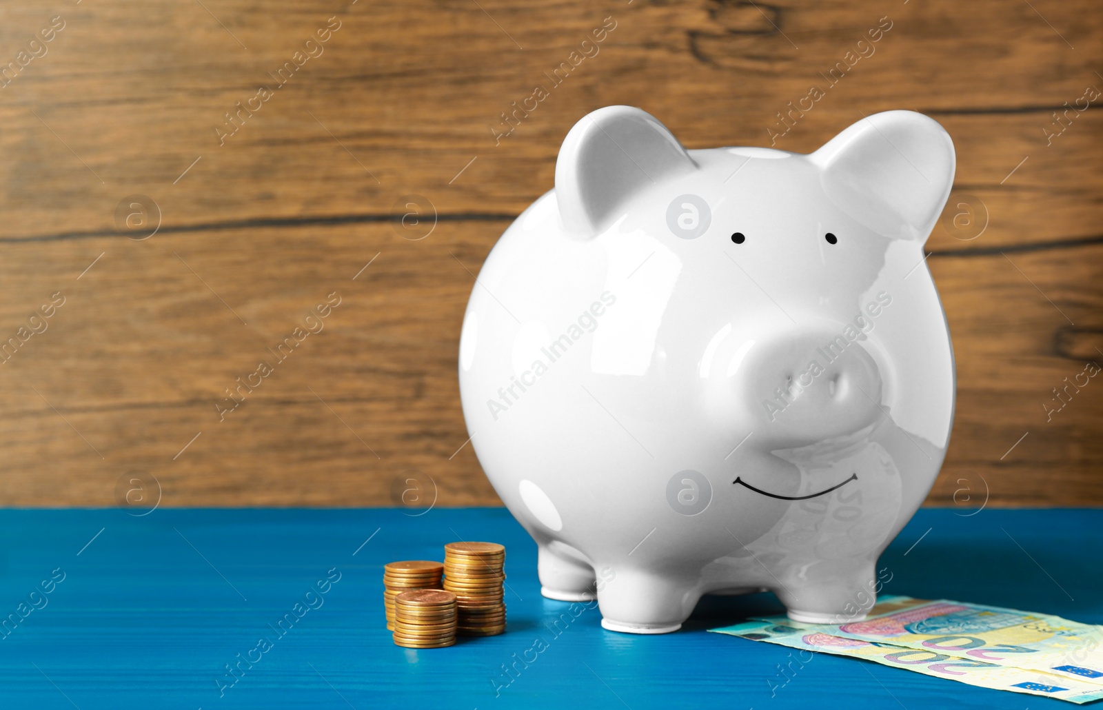 Photo of Ceramic piggy bank, banknotes and coins on blue wooden table. Space for text