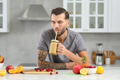Photo of Handsome man drinking delicious smoothie at white table in kitchen