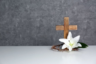 Photo of Wooden cross, crown of thorns and blossom lily on table against color background, space for text