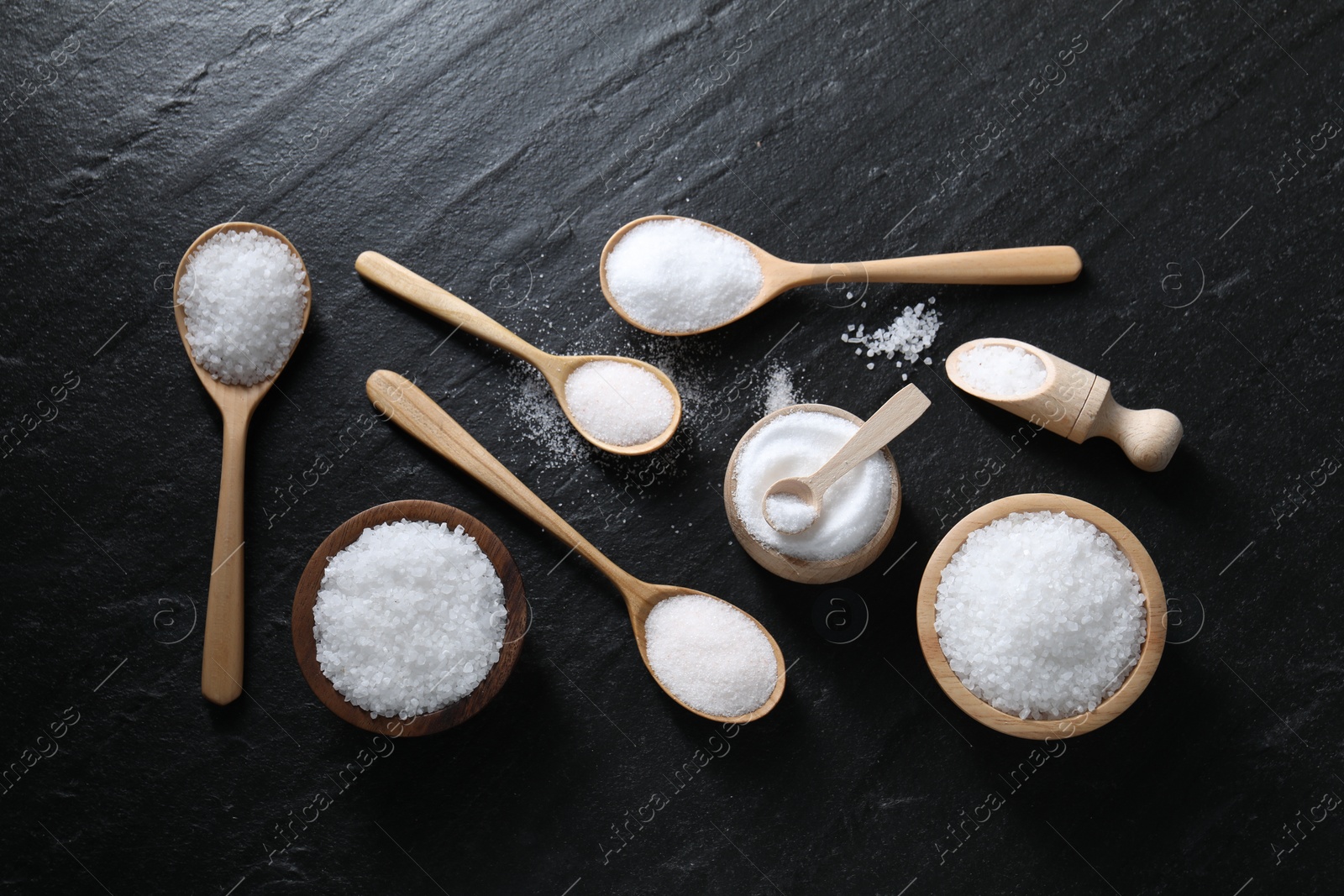 Photo of Organic white salt in bowls and spoons on black table, flat lay