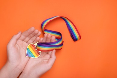 Photo of Woman holding rainbow ribbon with heart shaped pendant on orange background, top view and space for text. LGBT pride