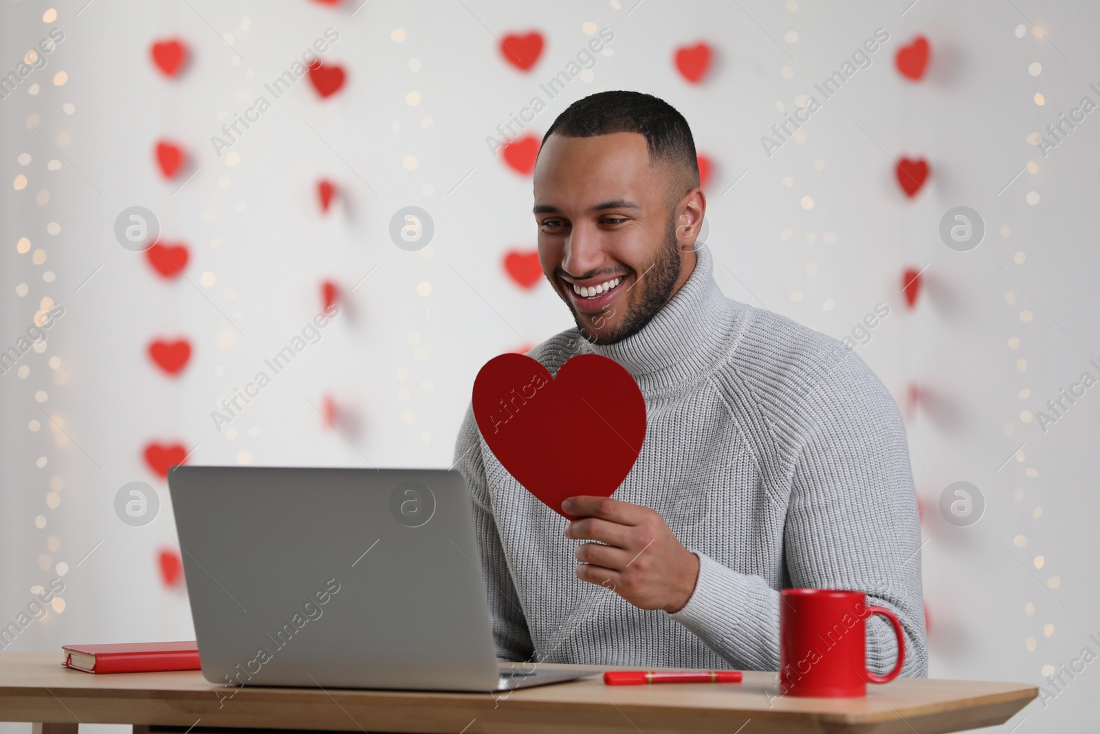 Photo of Valentine's day celebration in long distance relationship. Man holding red paper heart while having video chat with his girlfriend via laptop indoors