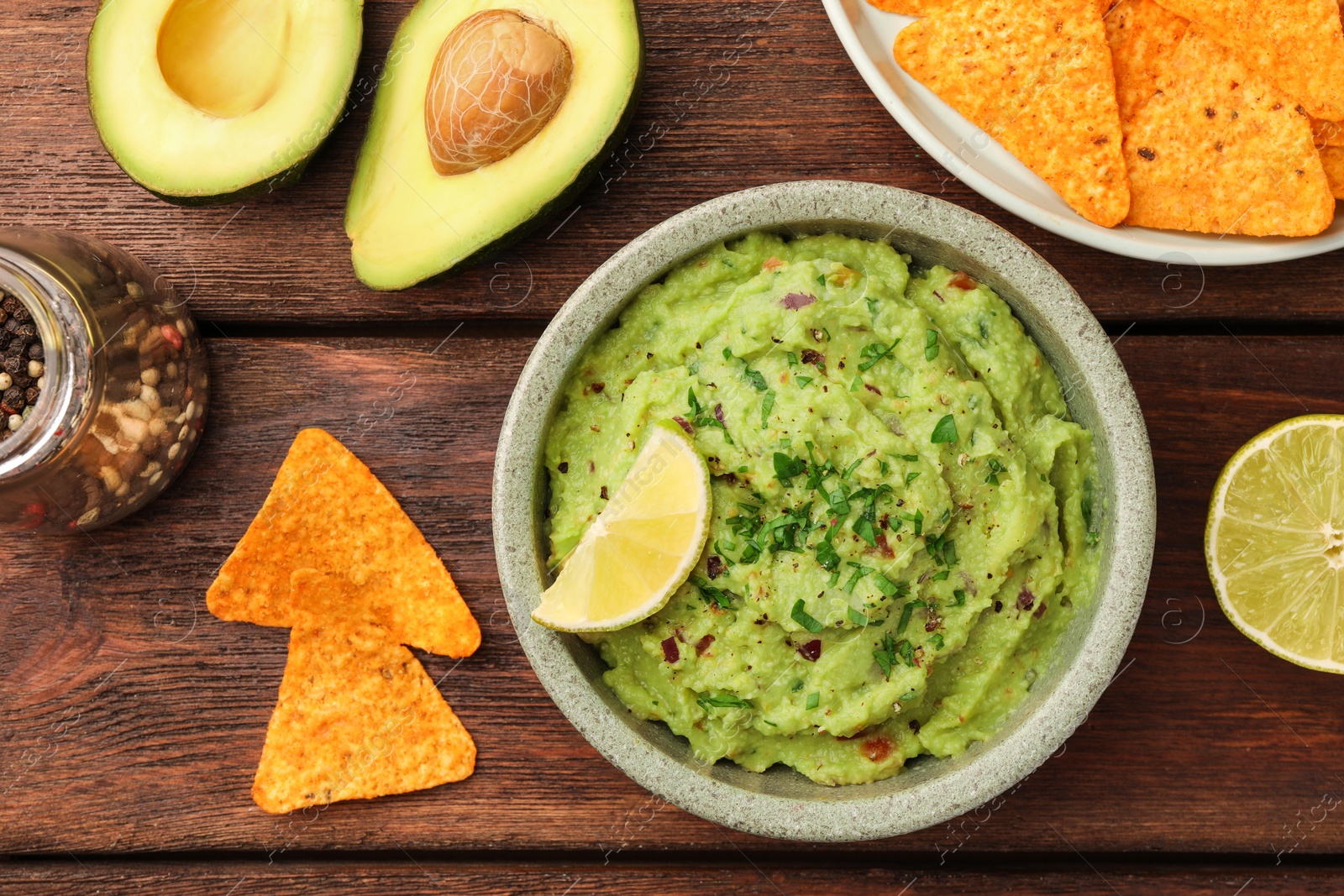 Photo of Bowl of delicious guacamole, lime and nachos chips on wooden table, flat lay