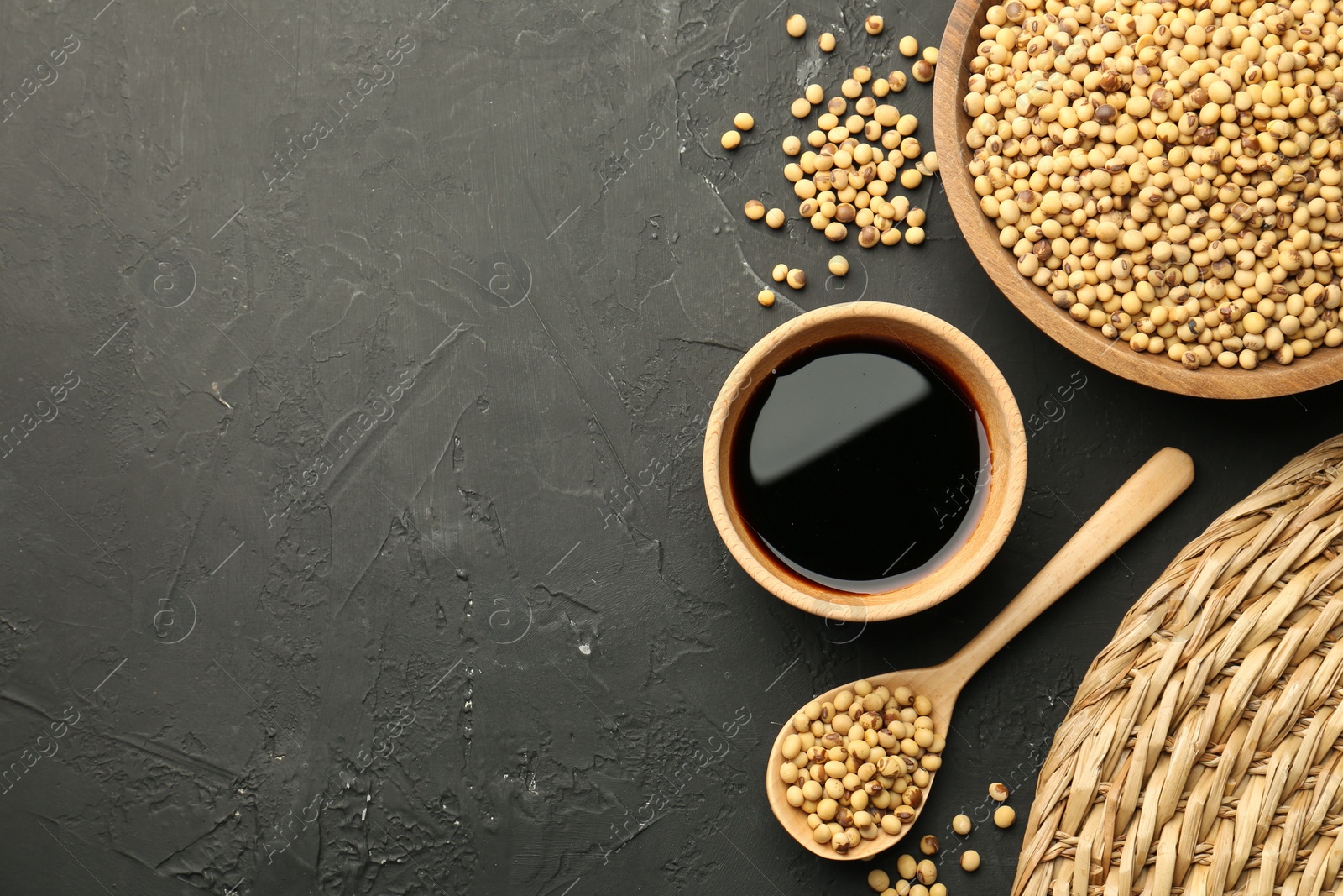 Photo of Soy sauce in bowl and soybeans on black textured table, flat lay. Space for text