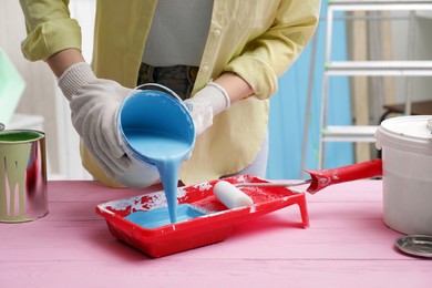 Photo of Woman pouring light blue paint from can into tray at pink wooden table indoors, closeup
