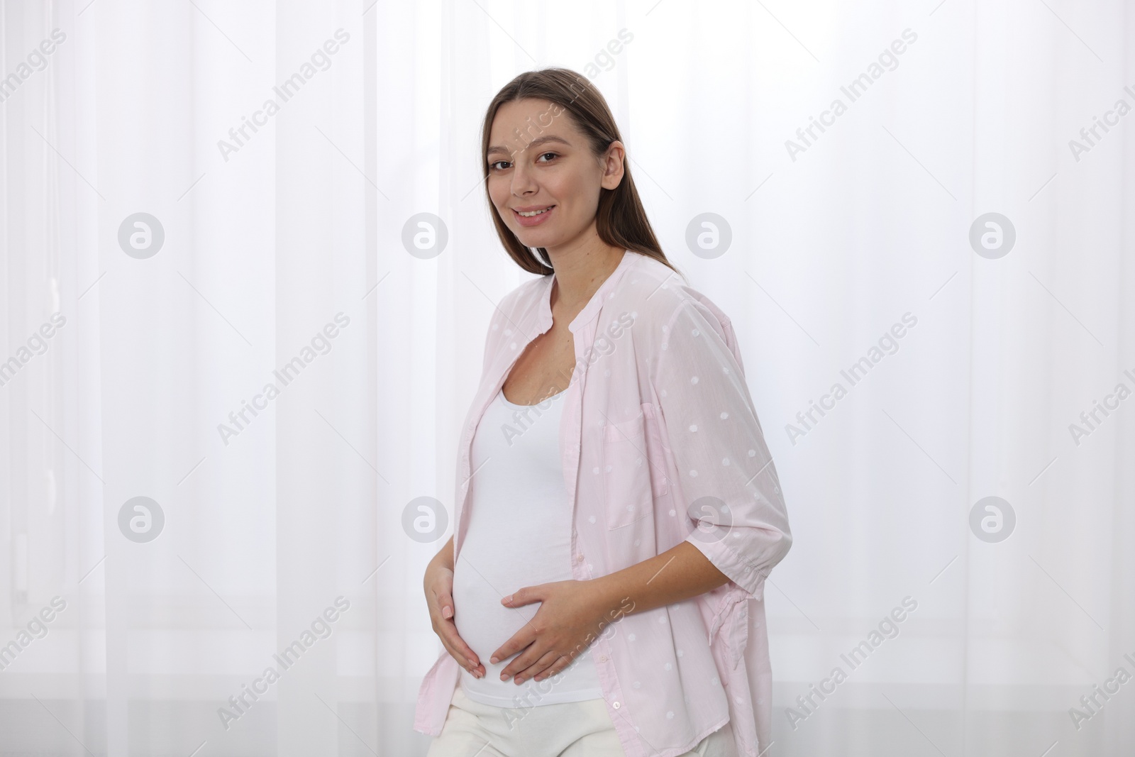 Photo of Beautiful pregnant woman in pink shirt near window indoors