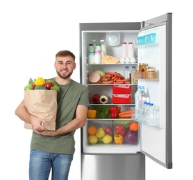 Photo of Young man with bag of groceries near open refrigerator on white background