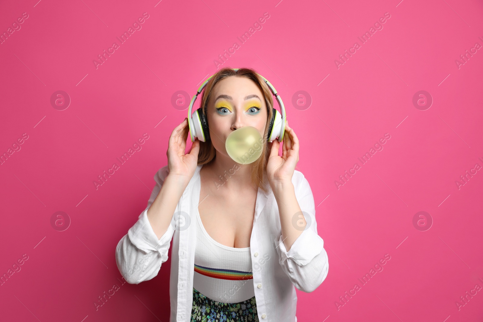 Photo of Fashionable young woman with bright makeup and headphones blowing bubblegum on pink background