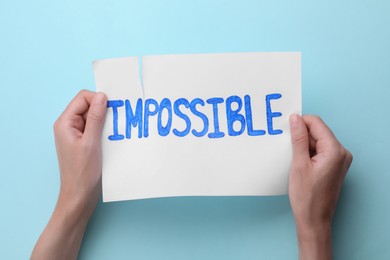 Photo of Motivation concept. Woman making word Possible from Impossible by tearing paper on light blue background, top view