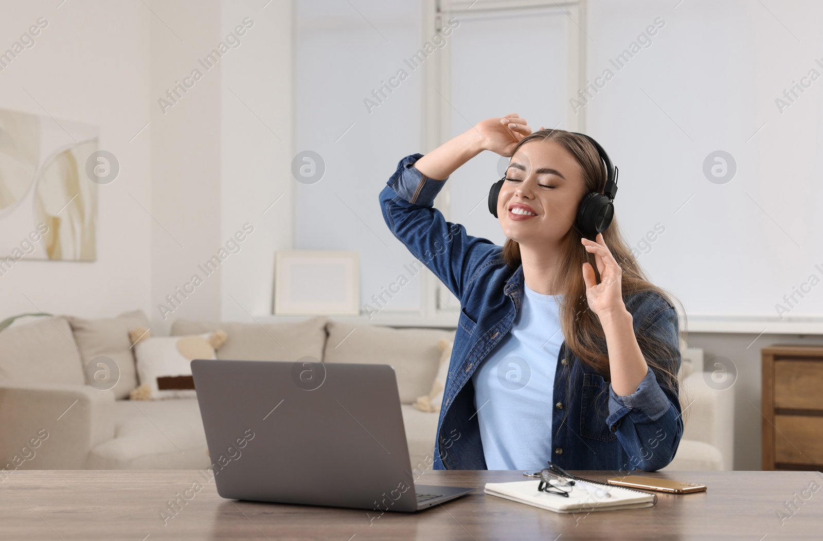 Photo of Happy woman with headphones listening to music near laptop at wooden table in room