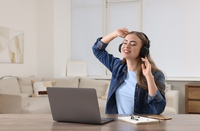 Happy woman with headphones listening to music near laptop at wooden table in room