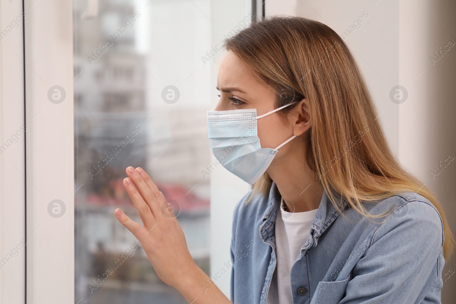 Photo of Sad woman in protective mask looking out of window indoors. Staying at home during coronavirus pandemic
