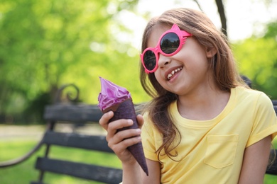 Cute little girl with delicious ice cream on bench in park, space for text