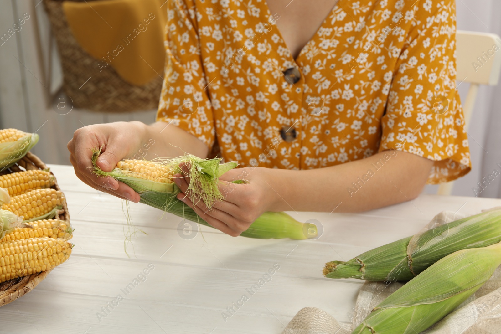 Photo of Woman husking corn cob at white wooden table, closeup