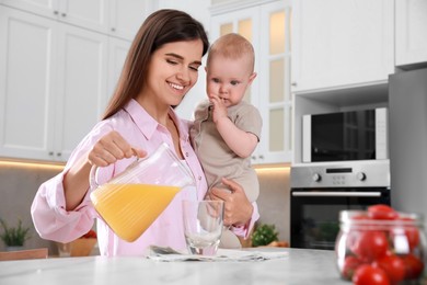 Mother holding her cute little baby while pouring juice into glass in kitchen, space for text