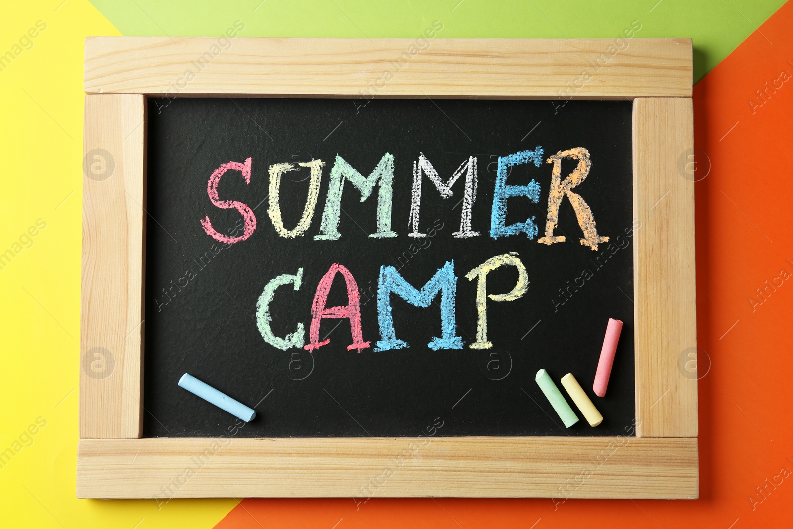 Photo of Small blackboard with text SUMMER CAMP and chalk sticks on color background, top view