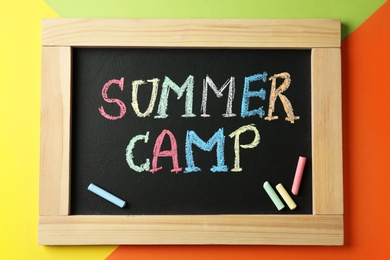 Small blackboard with text SUMMER CAMP and chalk sticks on color background, top view