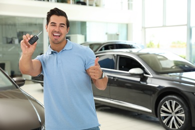 Happy young man with car key in modern auto dealership. Space for text